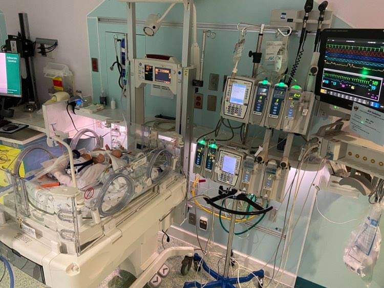 The image depicts a NICU hospital room, with a small incubator type bed, with IV poles and medical equipment surrounding it.